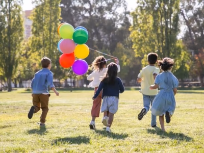 Grupo de niños corriendo por el césped con globos de colores en un parque público.
