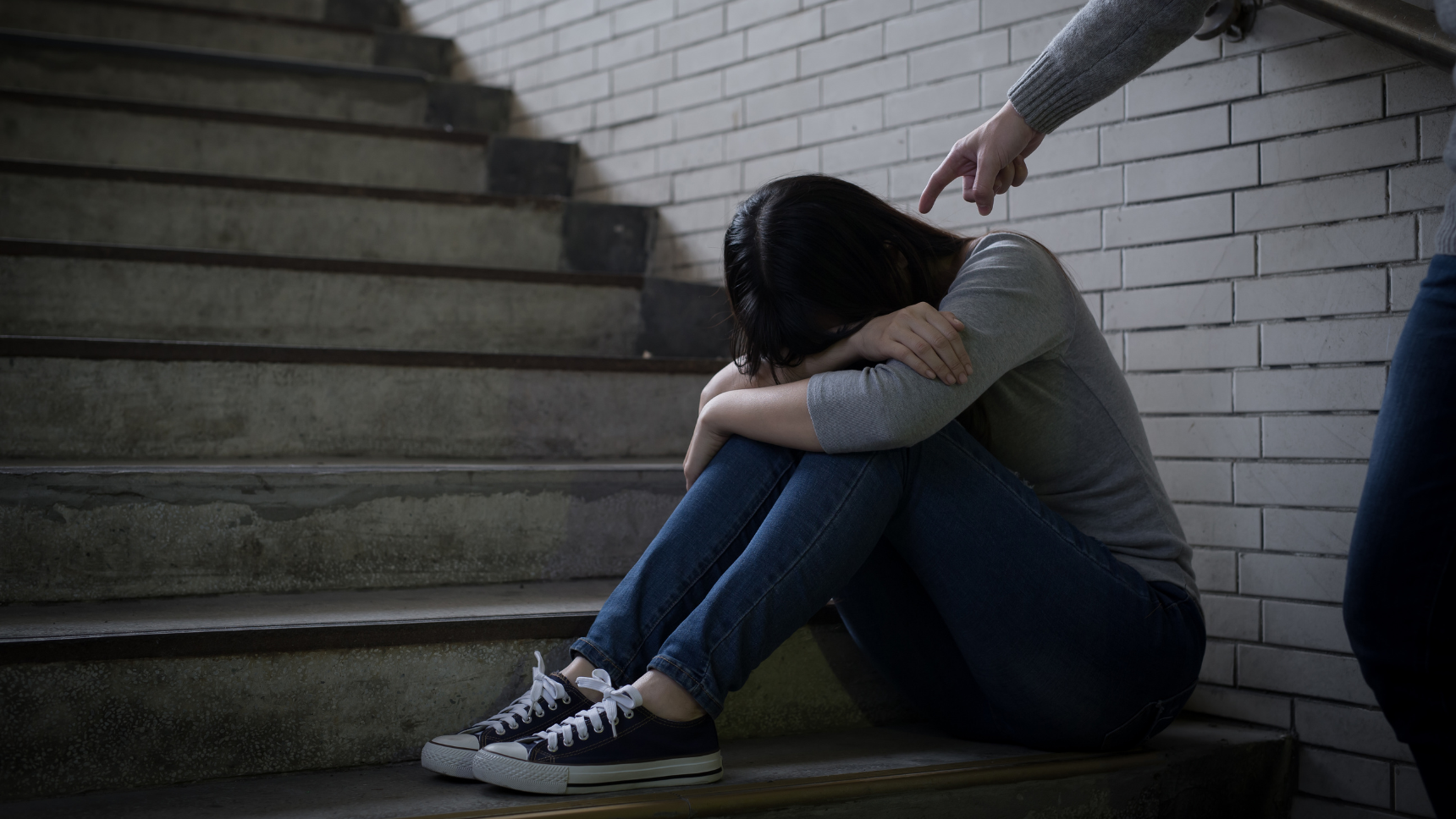 Adolescente víctima de bullying y acoso escolar sentada en unas escaleras mientras otra persona la señala