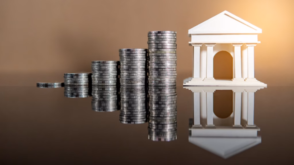 Maqueta de un banco junto a monedas apiladas que representan las cuentas bancarias en España de un extranjero fallecido.